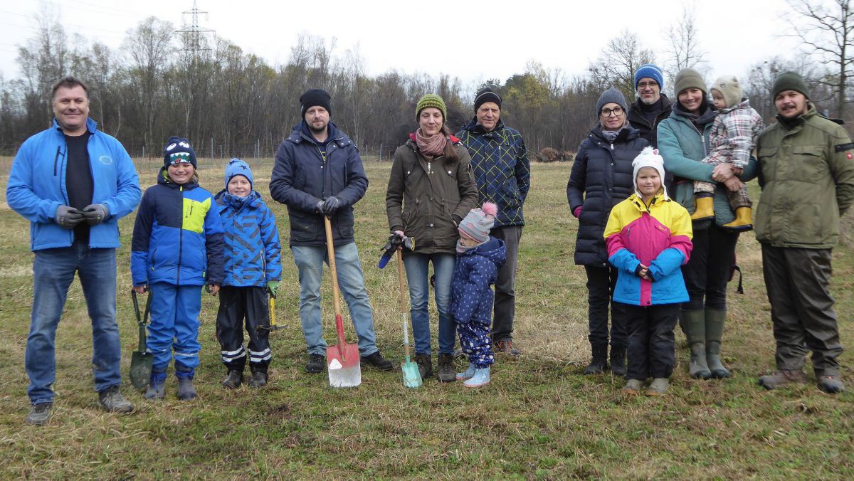 Gruppe von Menschen in winterlicher Kleidung steht auf einer Wiese, einige halten einen Spaten in der Hand. Im Hintergrund ist Wald zu erkennen.