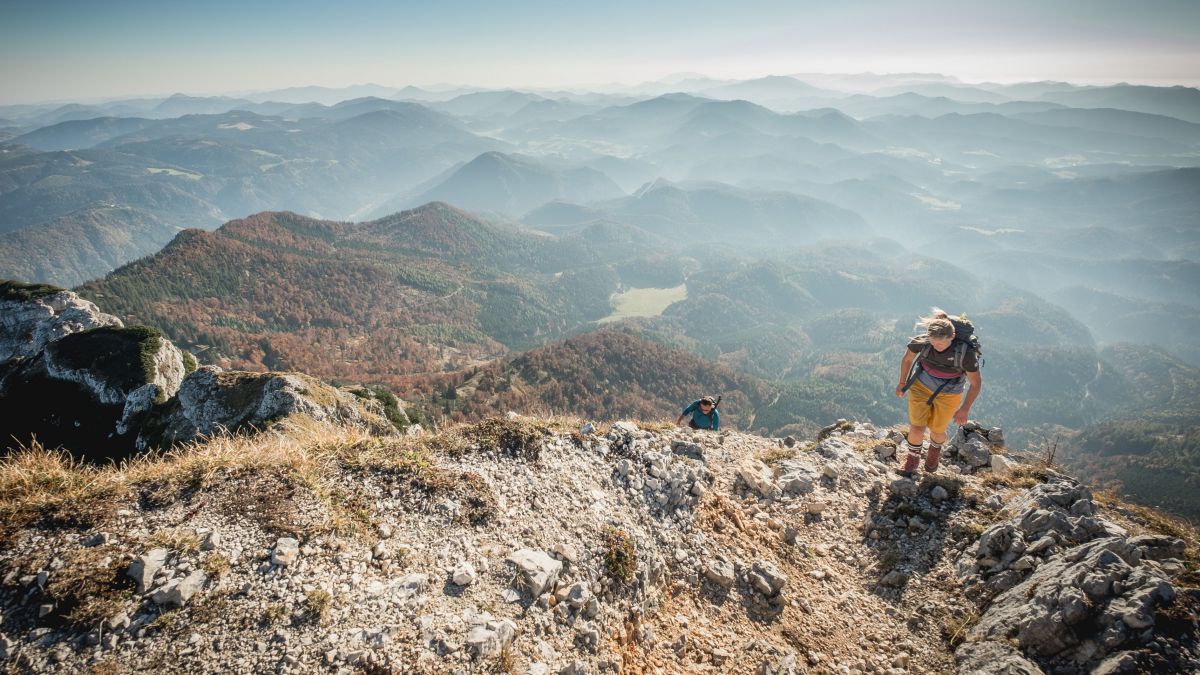 Person steigt auf einen Berg, im Hintergrund sieht man auf ein von Nebelschwaden umgebenes Gebirge