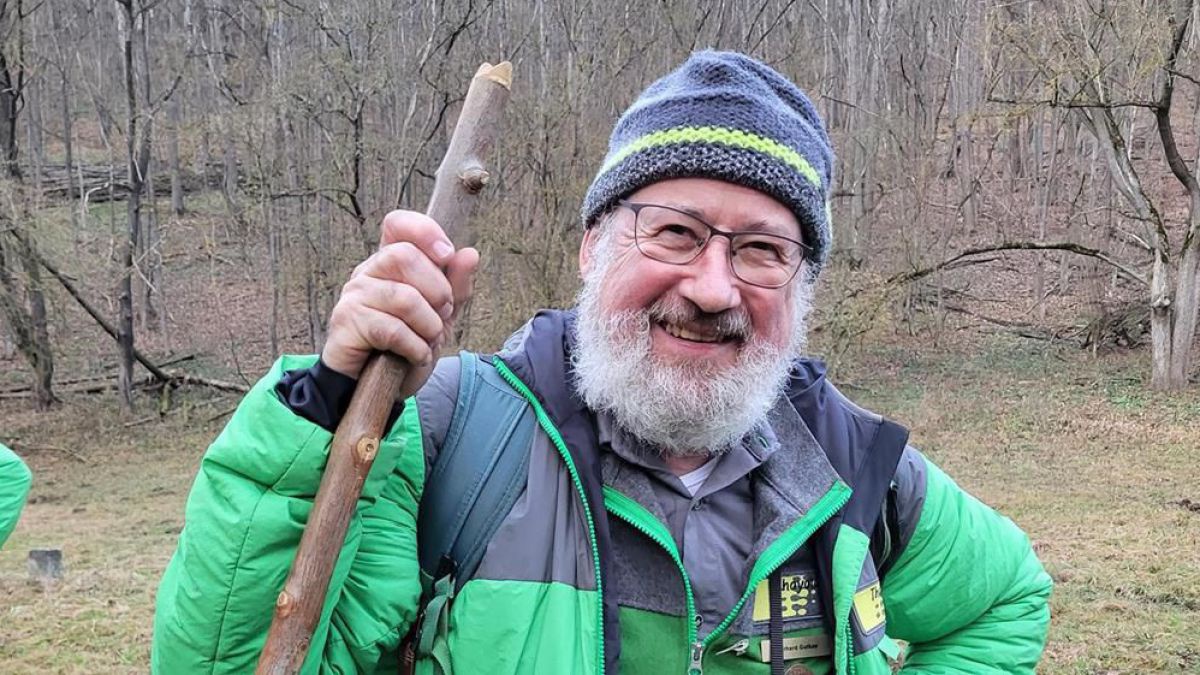 älterer Mann mit Bart und Brille mit grünem Anorak und Wollhaube lächelt in die Kamera, in der Hand hält er einen Holzstock.